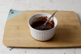 White ramekin of chocolate sauce with a small whisk in it, sitting on a bamboo cutting board with marble backdrop