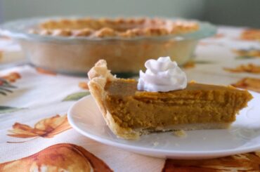 slice of pumpkin pie on a white plate with a pie in a glass dish behind it