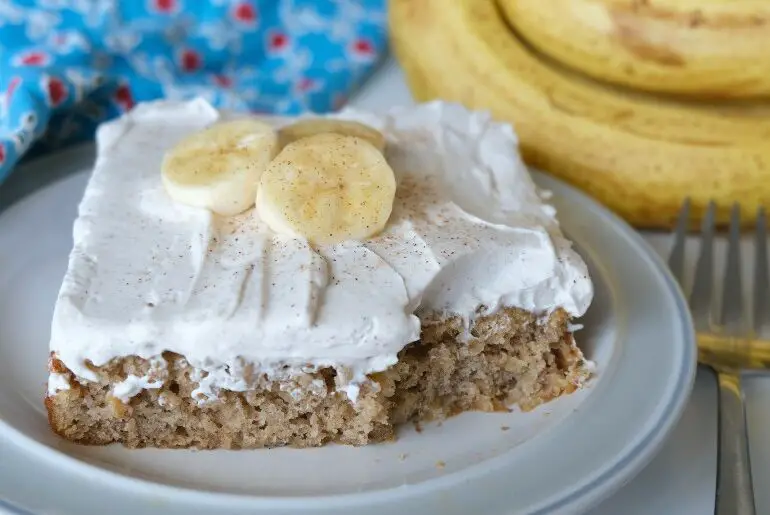 square banana cake with a bite taken out is on a pale green plate. The cake is golden brown with white, fluffy frosting and 3 banana slices on top. There are several bananas and a blue floral towel in the background.