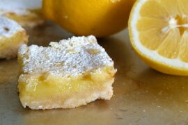 A single lemon bar placed on a sheet pan, with lemons visible in the background. The lemon bar is square-shaped with a golden brown crust and a vibrant yellow lemon filling that is visible in the center.