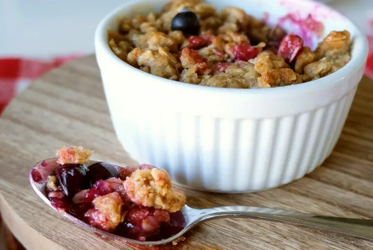 a spoonful of blueberry crisp on a spoon sits on a wooden surface with a blueberry crisp in a white ramekin in the background