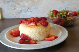 small, round, pale yellow cake on a plain plate topped with sliced strawberries. A small bowl of strawberries is in the background.