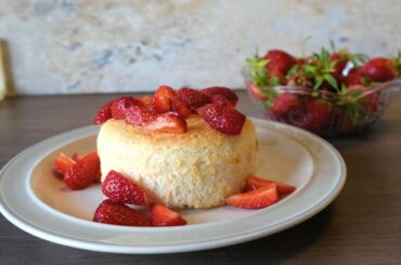 small, round, pale yellow cake on a plain plate topped with sliced strawberries. A small bowl of strawberries is in the background.