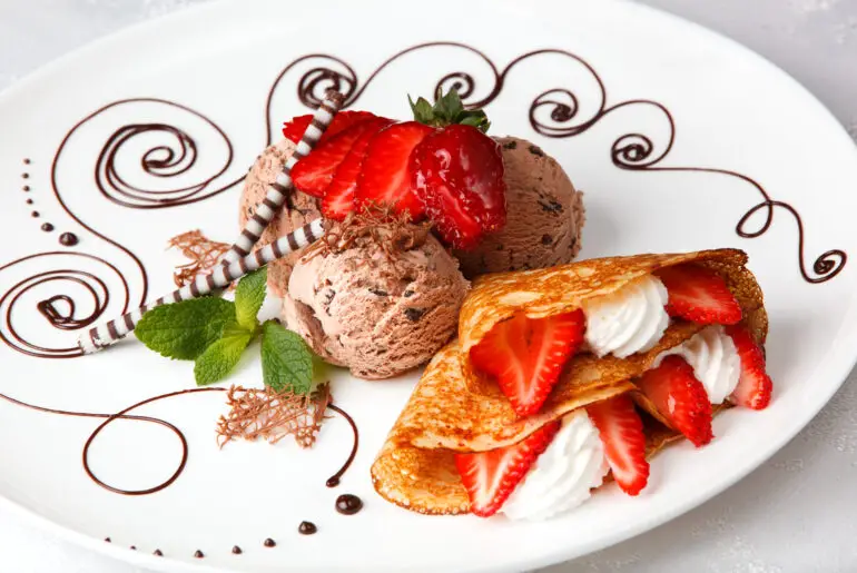 two crepes filled with whipped cream and strawberries next to several scoops of chocolate ice cream all on a white plate surrounded by thin swirls of chocolate sauce in a decorative pattern.