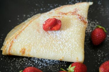 lightly golden crepe folded into a triangle on black plate. The crepe is dusted with powdered sugar and surrounded by red strawberries