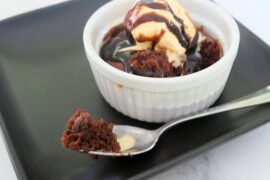 A brownie in a white ramekin sits on a black plate. The brownie is topped with ice cream and chocolate sauce. A silver spoon with a bite of brownie rests on the plate in front of the ramekin.