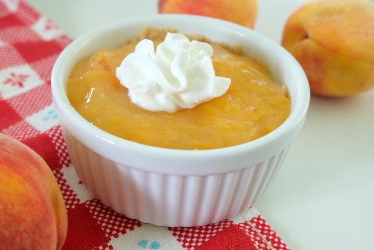 image features a mini peach pie in a white ramekin on a red and white checkered cloth. There are several peaches around the pie and the pie is topped with a small dollop of whipped cream.